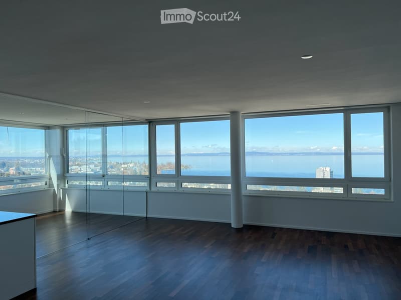 Empty room with large windows offering a scenic view of water and distant cityscape, featuring wooden flooring and modern lighting.