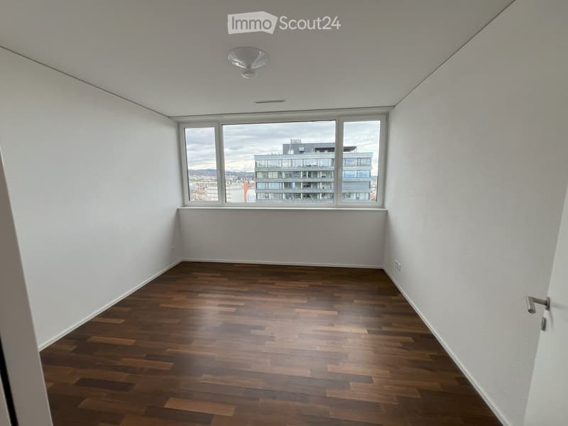 Empty room with wooden flooring, white walls, and large windows offering a view of a modern building.
