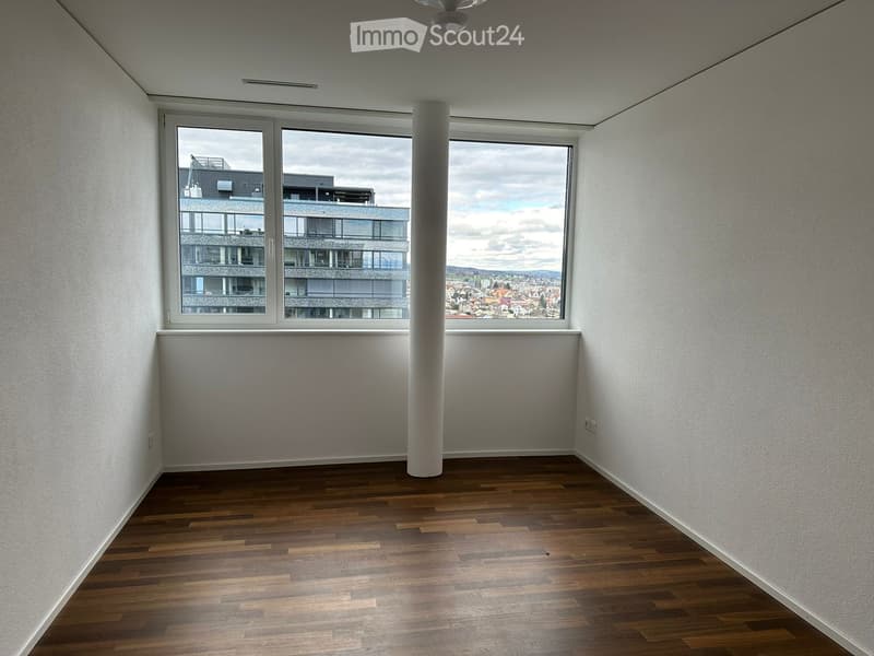 Empty room with wooden flooring, white walls, and large windows offering a view of a modern building and cityscape.