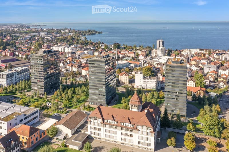 Aerial view of a cityscape with a mix of residential and commercial buildings, surrounded by greenery and a body of water in the background.
