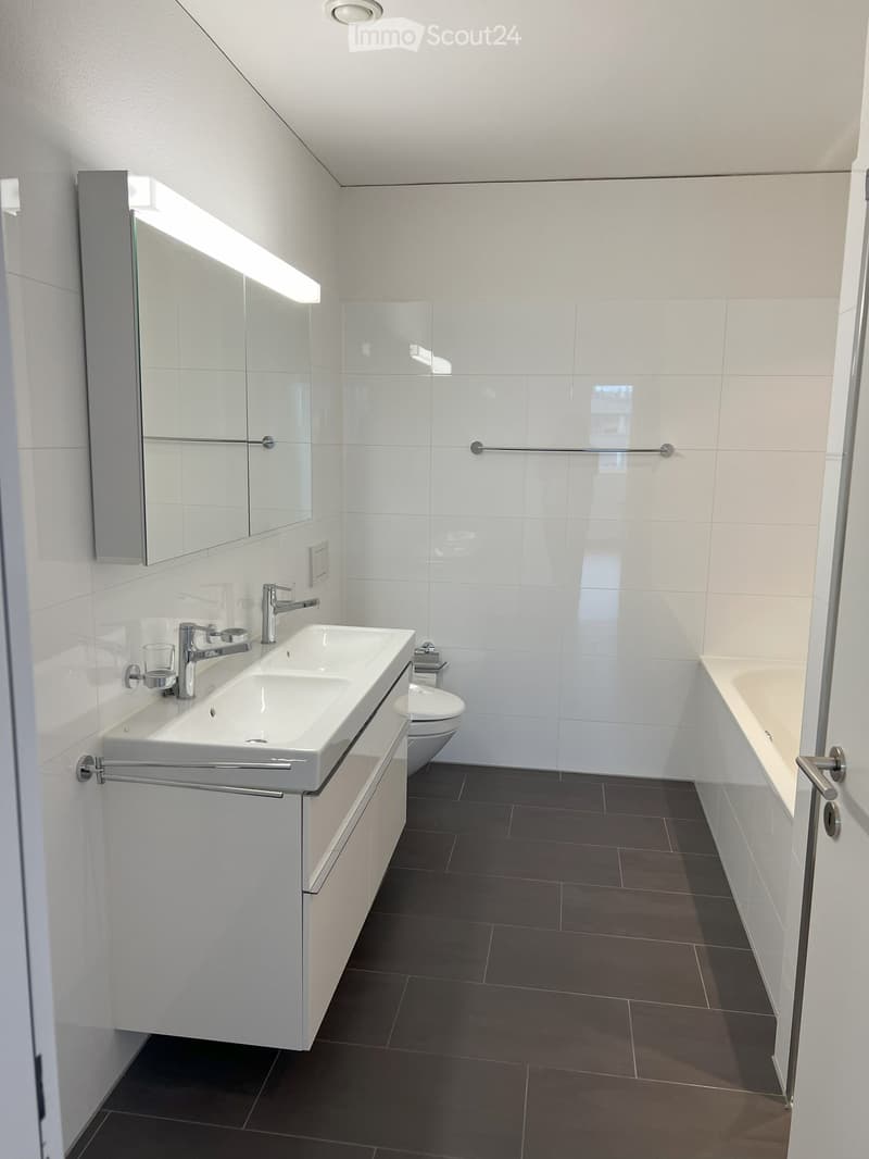 Modern bathroom with white tiles, dark floor tiles, double sink, mirror, toilet, and bathtub.