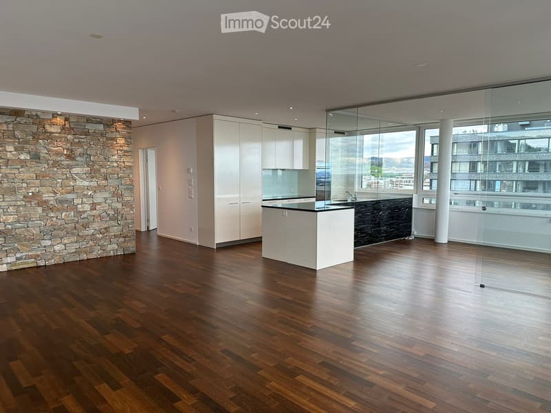 Modern kitchen with light-colored cabinets, a central island, and a dark countertop, featuring a stone accent wall and large windows with a city view.