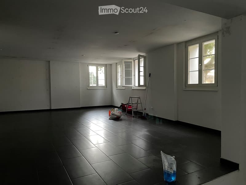 Empty room with dark tiled floor, white walls, multiple windows, and cleaning supplies on the floor.