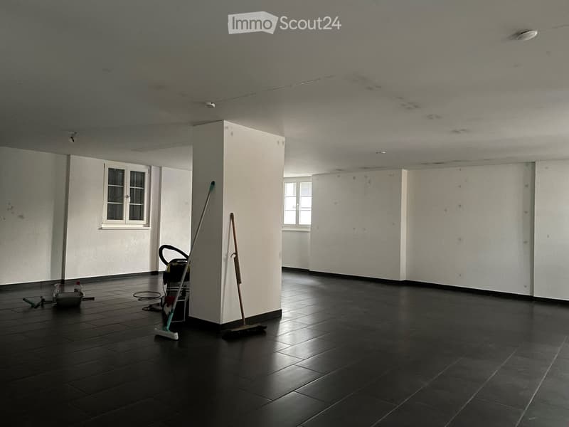 Empty room with bare walls, dark tiled floor, and a few cleaning tools placed near the entrance.