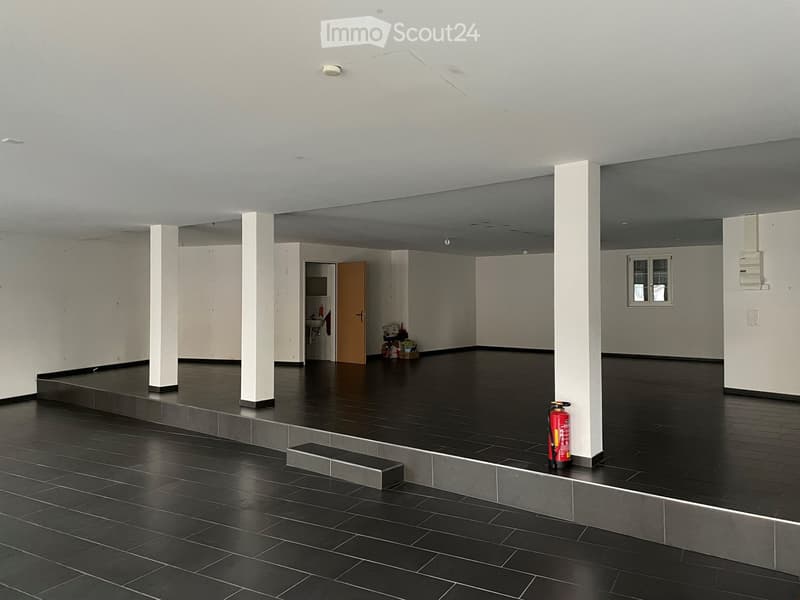 Empty room with dark tiled floor, white walls, and white pillars. A small bathroom area is visible in the background, and a red fire extinguisher is placed near a pillar.