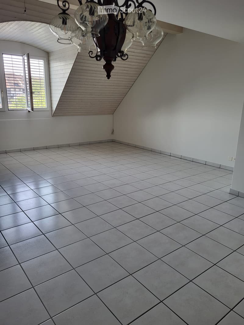 Empty room with white walls, tiled floor, and a chandelier hanging from the ceiling. A window with blinds is visible on one wall.