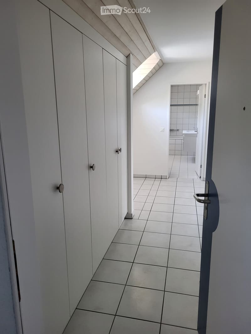 A narrow, tiled hallway with white walls, leading to a small bathroom with a sink and bathtub. The hallway features built-in white cabinets on one side and a door at the end.