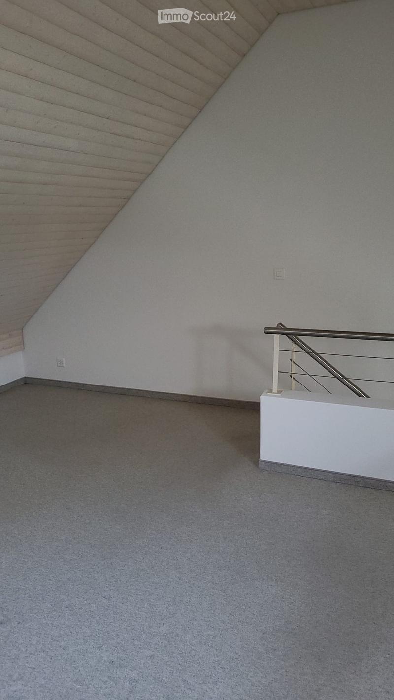 Empty room with grey carpeted floor, white walls, and a wooden slanted ceiling. A white staircase railing is visible on the right side.
