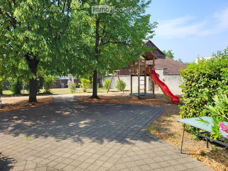 Outdoor area with a paved walkway, large trees, a wooden playhouse with a red slide, and a small table with flowers.