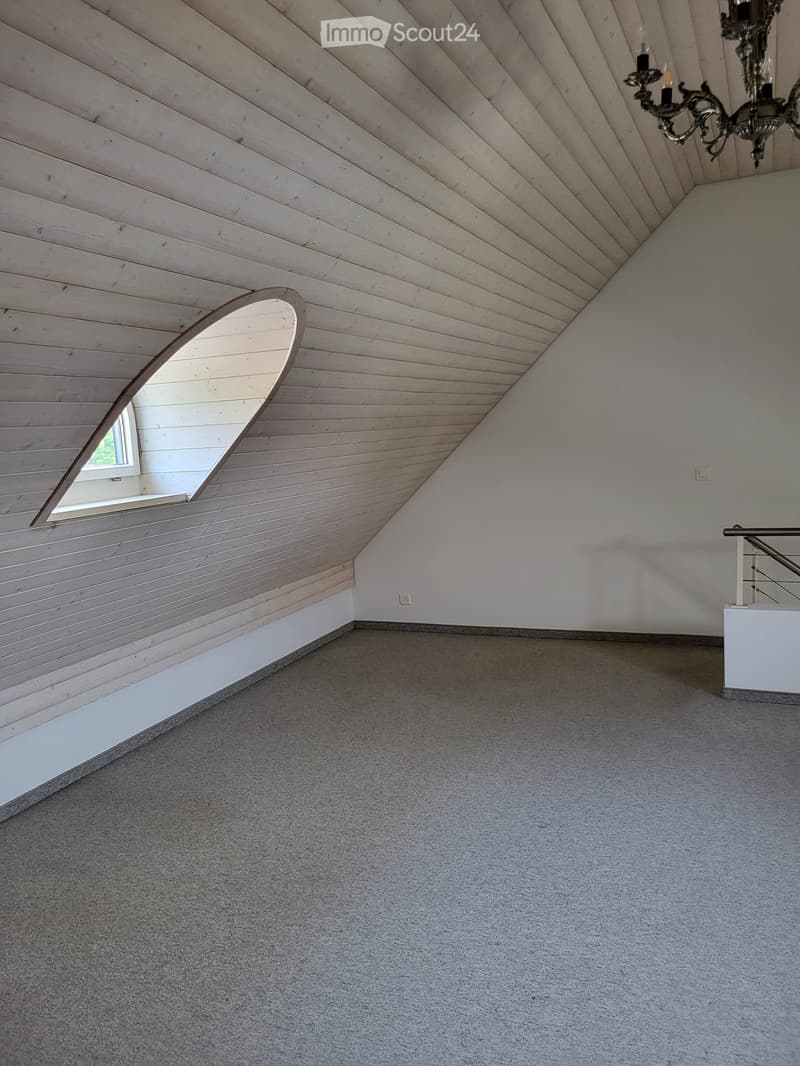 Empty attic space with wooden slatted ceiling, grey carpeted floor, white walls, and a small window with a wooden frame.