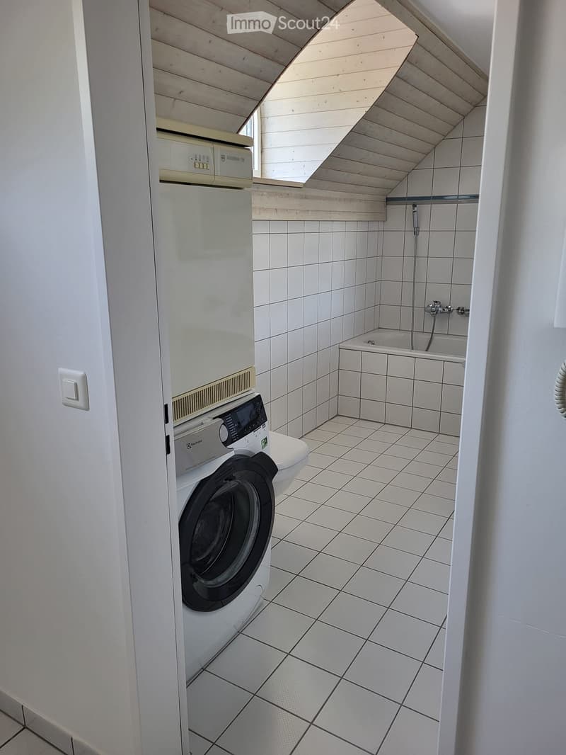 Compact bathroom with white tiled walls and floor, featuring a washing machine, bathtub, and faucet.