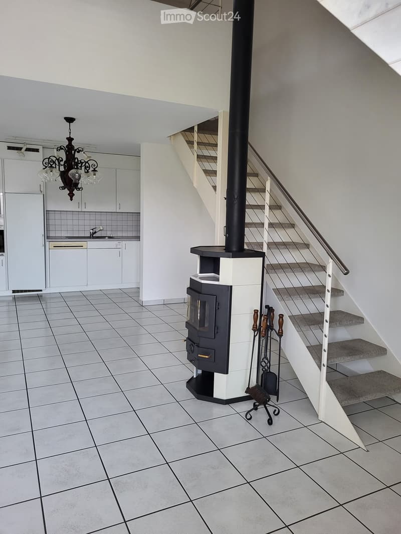Modern interior with a staircase, fireplace, and kitchen area visible. The space features tiled flooring, white walls, and a chandelier hanging from the ceiling.