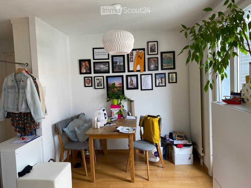 A cozy, well-lit room with a wooden table, two chairs, a sewing machine, a plant, and several picture frames on the wall. A large window with a curtain lets in natural light, and a coat rack with jackets is visible on the left side.