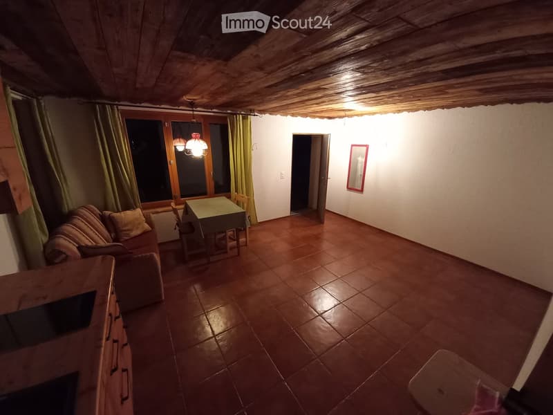 A cozy living room with a wooden ceiling, tiled floor, and large window with curtains. Features a sofa, small dining table with chairs, and a mirror on the wall.