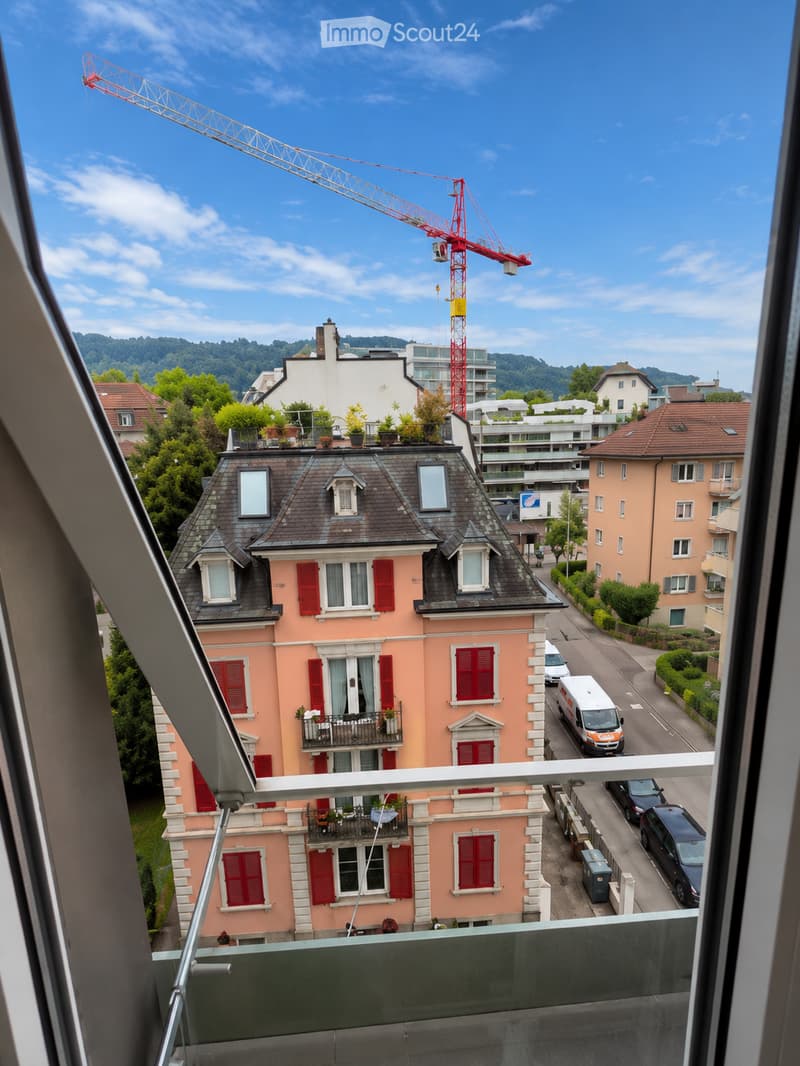 View from a balcony showing a multi-story residential building with pink facade, red shutters, and multiple windows. The building has a dark roof with dormer windows and balconies with potted plants. A construction crane is visible in the background, alon