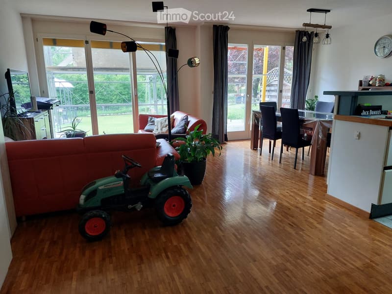 Open-plan living and dining area with wooden flooring, large windows, a red sofa, a toy tractor, a potted plant, a dining table with chairs, and a floor lamp.