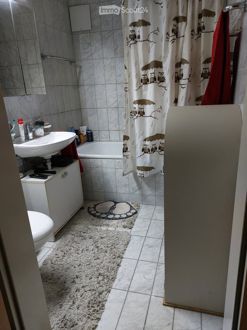 Bathroom with white tiles, a sink, bathtub, toilet, and a shower curtain with owl and umbrella designs. Includes a fluffy white rug and a smaller patterned rug.