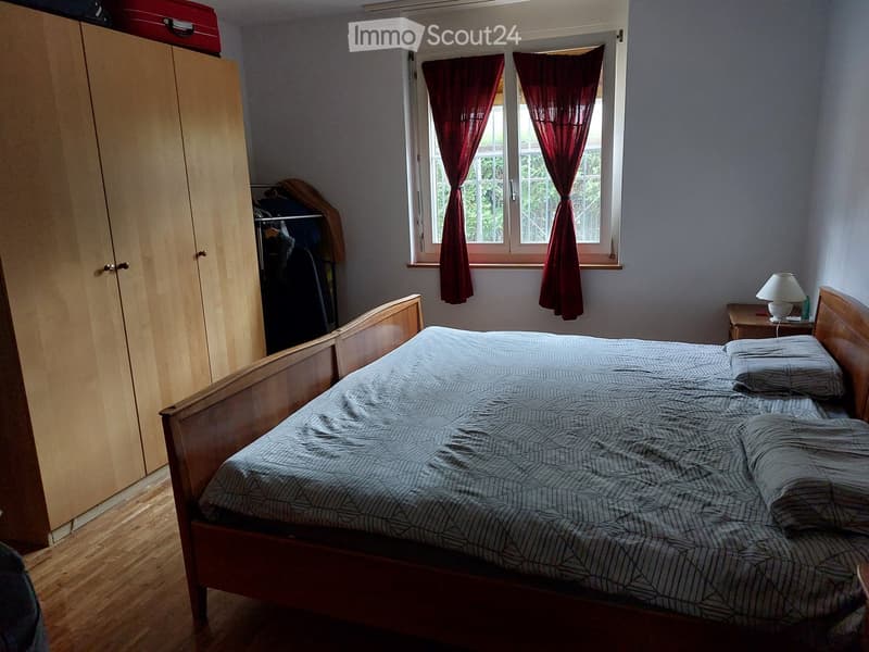 Bedroom with a wooden bed, nightstand, lamp, and wardrobe. The room has wooden flooring and a window with red curtains.