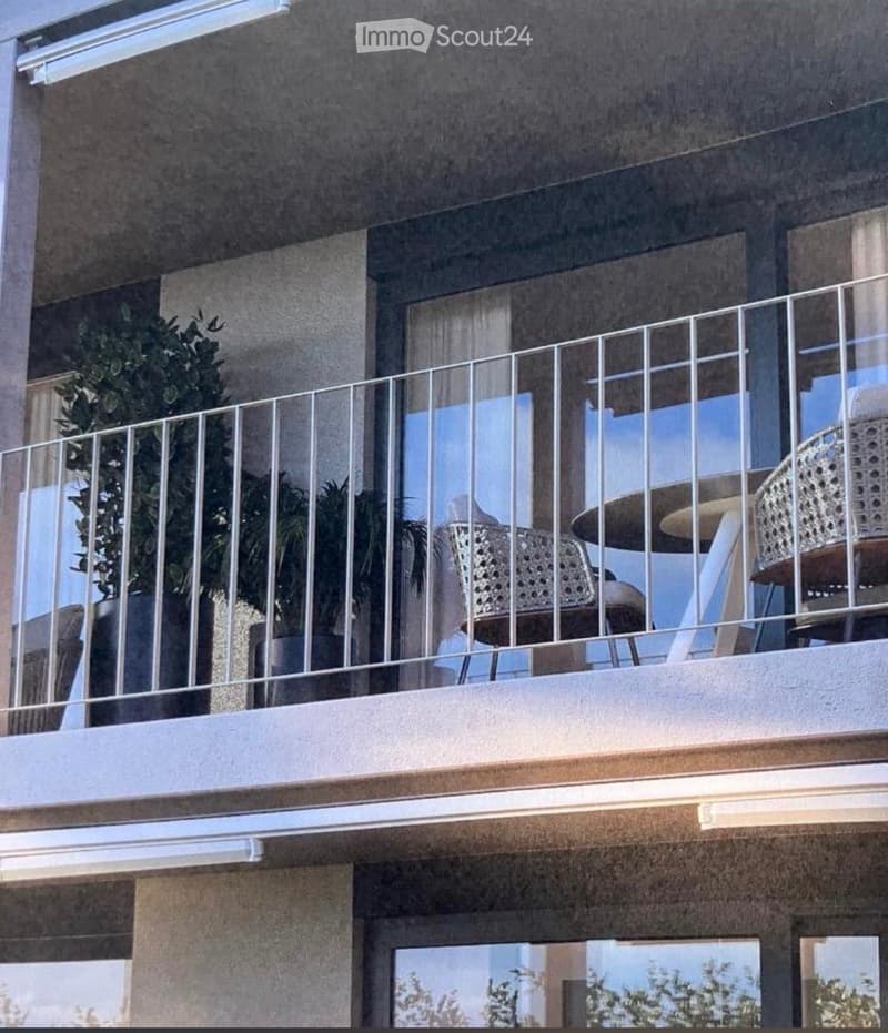 Balcony with metal railing, two chairs, and a small table, with potted plants visible in the background.