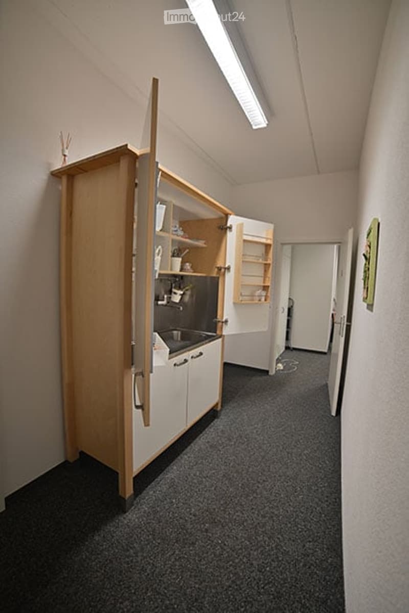 A small, furnished kitchen area with a wooden cabinet, sink, and shelves. The space is clean and organized with a gray carpeted floor and white walls.