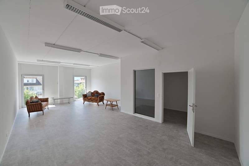 Bright, minimalist room with white walls and concrete flooring, featuring a sofa, small table, and chairs near large windows and a glass door leading to an outdoor area.