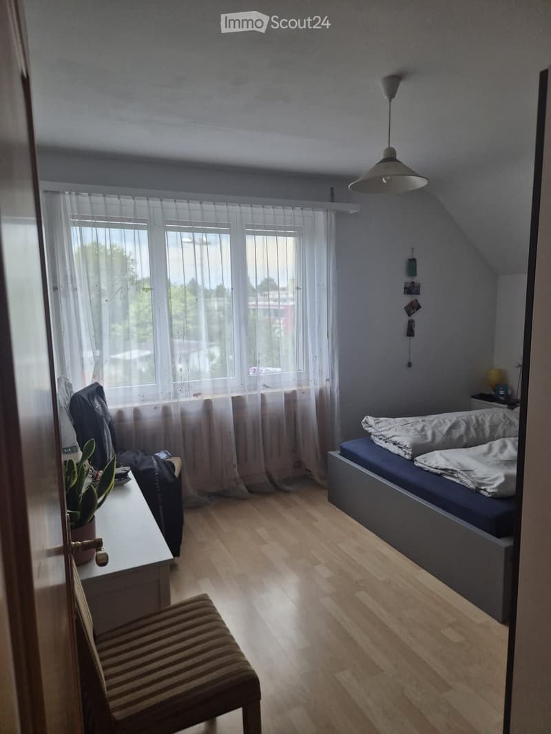 Bright bedroom with wooden flooring, large windows with sheer curtains, a bed with pillows and blankets, a chair, a small table with a potted plant, and a hanging light fixture.