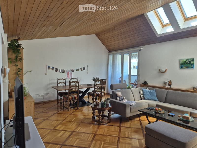 A cozy living room with a grey sectional sofa, coffee table, dining table with chairs, and a TV screen. The room features wooden flooring and a slanted wooden ceiling with skylights.
