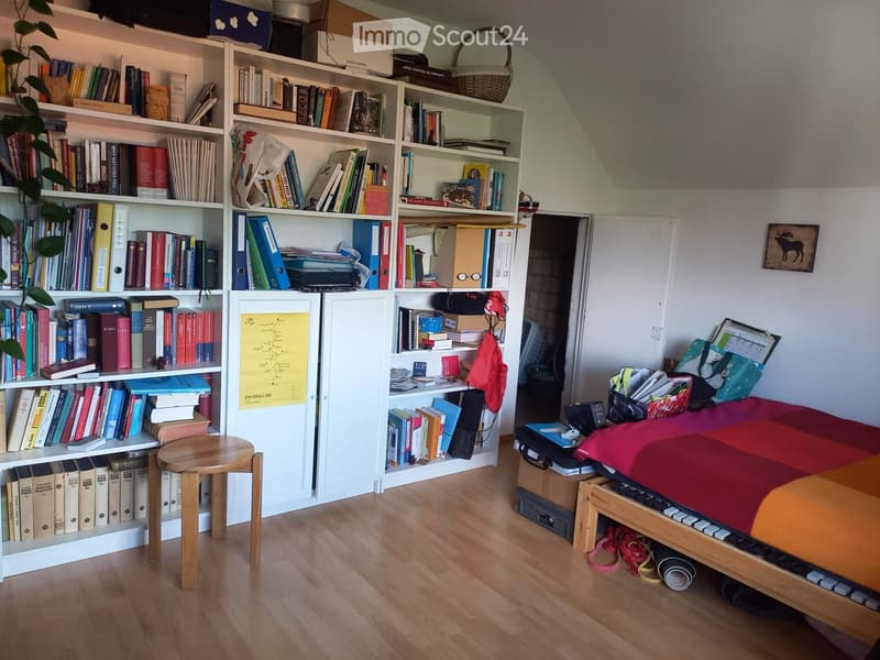 A cozy bedroom with a wooden bed, a small wooden stool, a large bookshelf filled with books, and a laptop on the bed. A plant is placed on the bookshelf, and a map is visible on the wall.