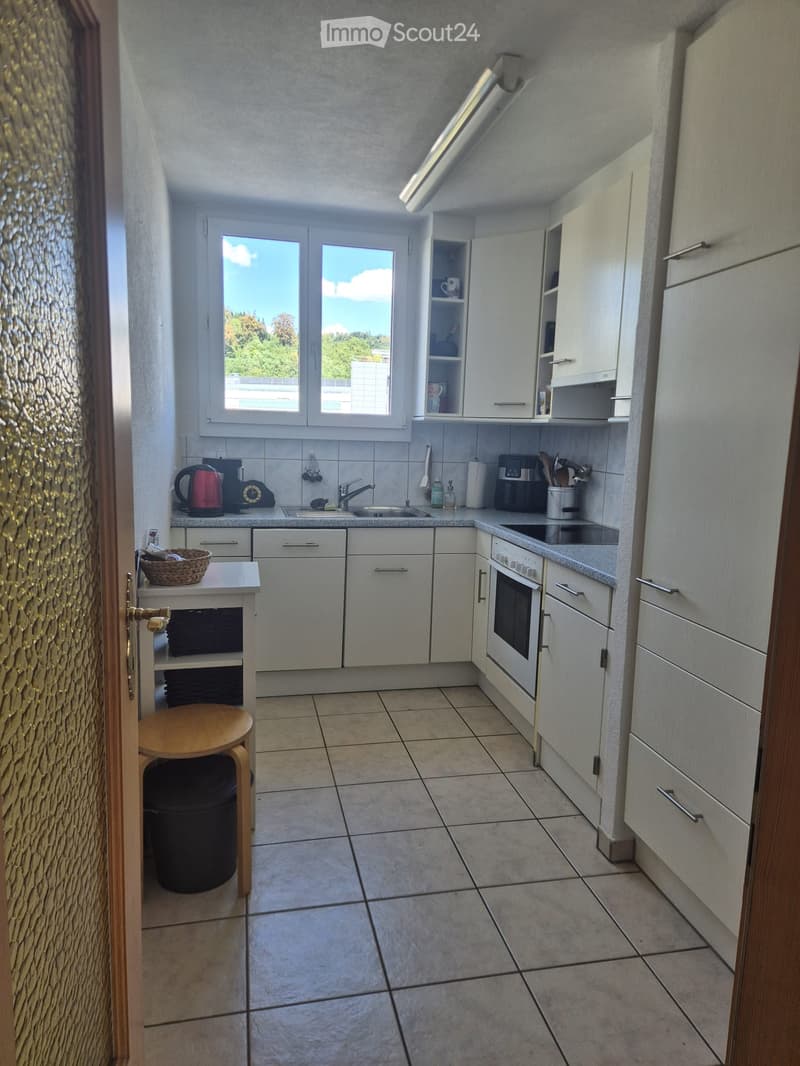 Modern kitchen with white cabinets, tiled floor, and various appliances including a coffee maker, kettle, and oven. A small wooden stool and a basket are placed near the entrance.