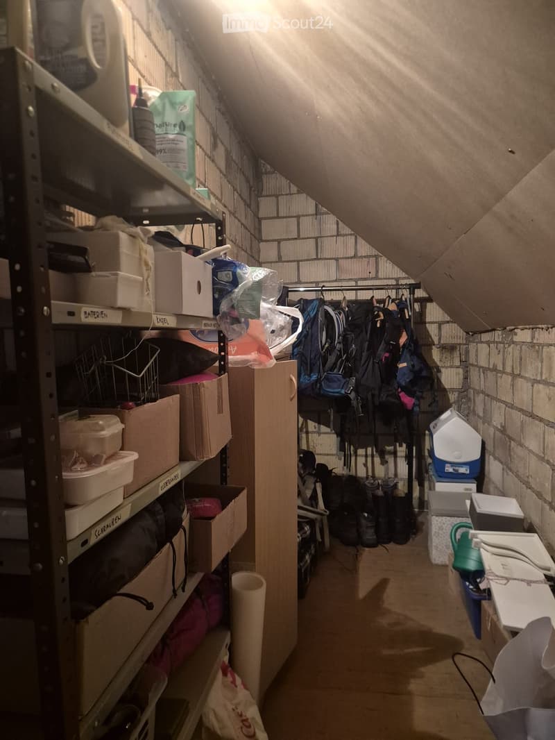 A narrow, slanted attic corridor with metal shelving units on one side and a clothing rack on the other. Shelves contain boxes, bags, and miscellaneous items, while the clothing rack holds jackets and bags.