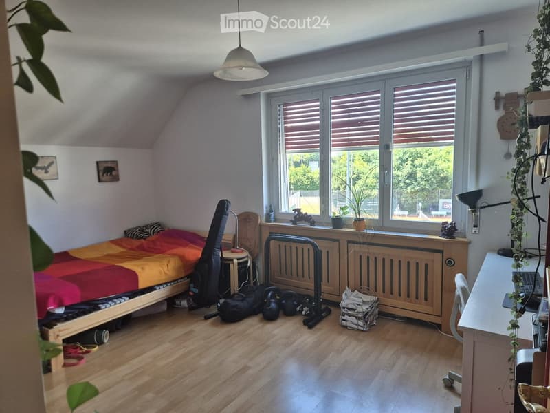 A cozy bedroom with a bed, desk, chair, and radiator. The room features wooden flooring, large windows with blinds, and decorative items including potted plants and picture frames.