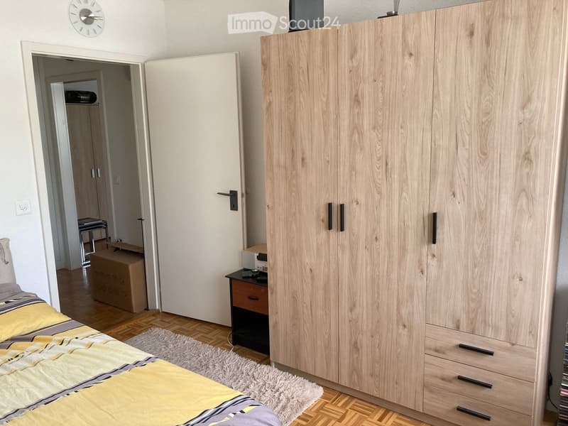 A furnished bedroom with a bed, wardrobe, nightstand, table, chair, and a clock on the wall.