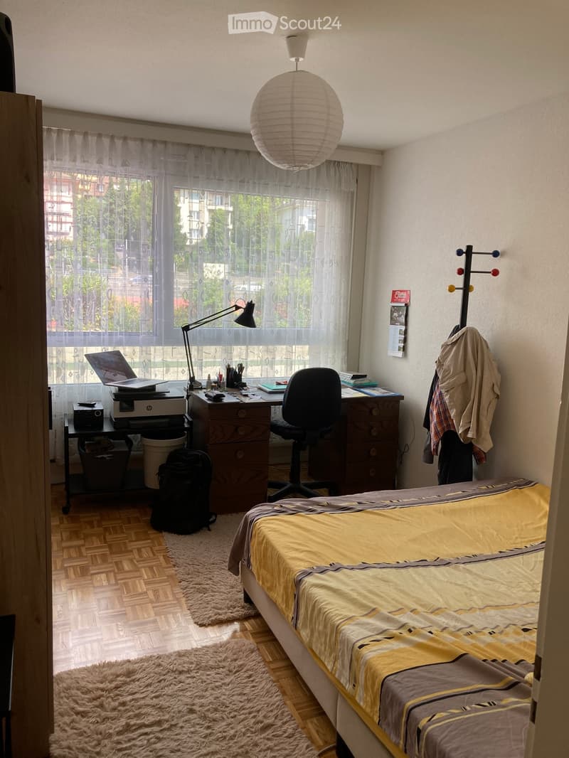 Cozy bedroom with a single bed, wooden desk, chair, laptop, printer, and a coat rack with clothes hanging. The room features a large window with sheer curtains, a round paper lantern light fixture, and a fluffy rug on the floor.