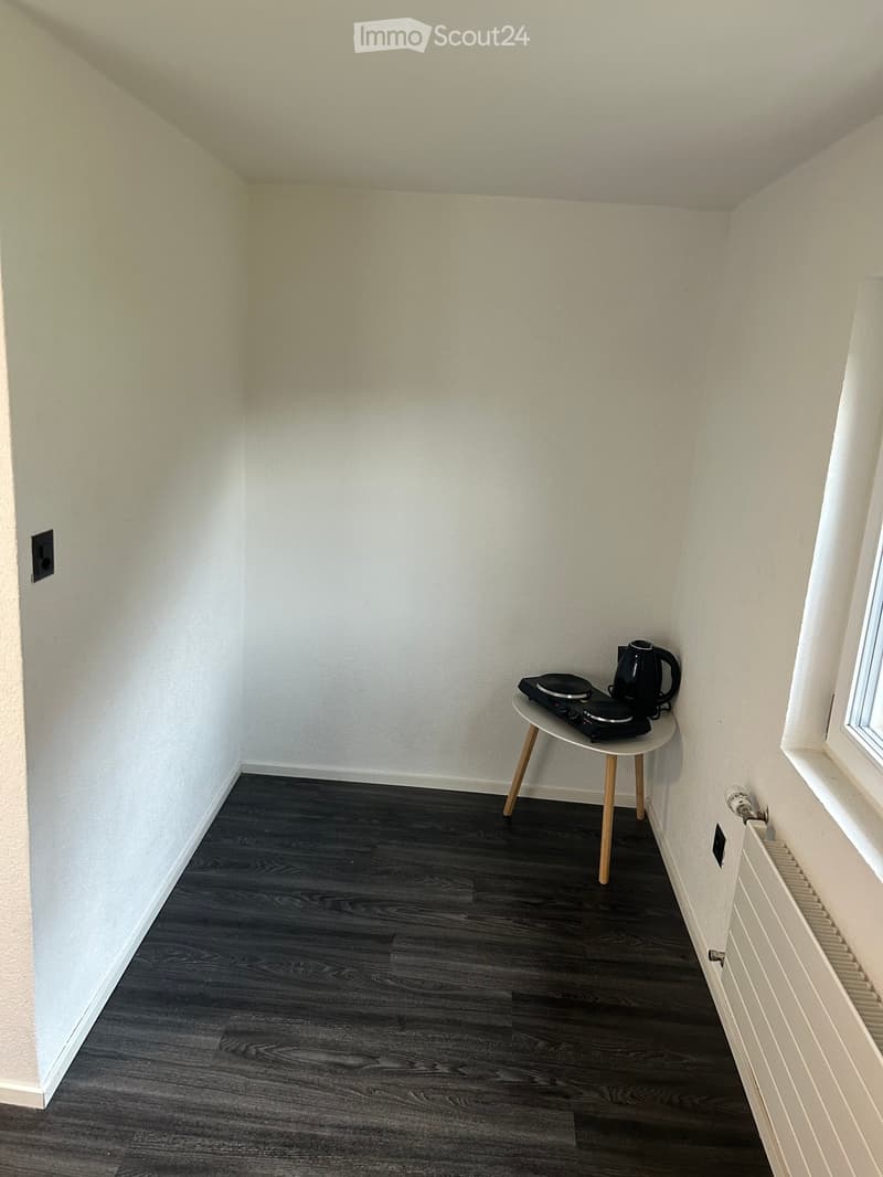 Small room with dark wooden flooring, white walls, a small round table with an induction cooker and kettle, and a radiator under a window.