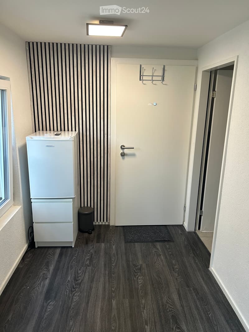 A small, furnished room with dark wooden flooring, a white refrigerator, a trash can, a door with a hook rack, and a small rug near the entrance.