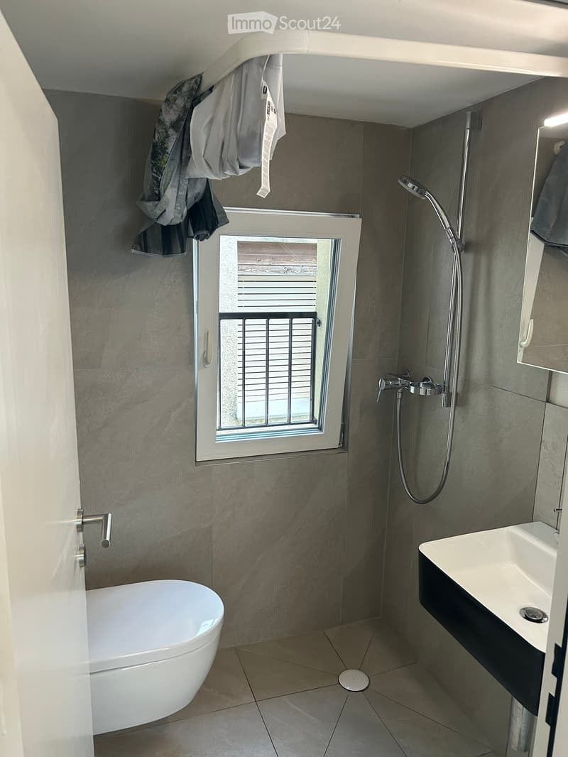 Modern bathroom with a white bathtub, a sink with a faucet, a showerhead, and a window with blinds. The walls are tiled in a neutral tone, and there is a small shelf with folded towels.