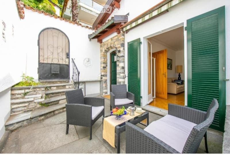 A cozy outdoor terrace with wicker chairs, a small table with a fruit bowl, and a view of the building entrance.