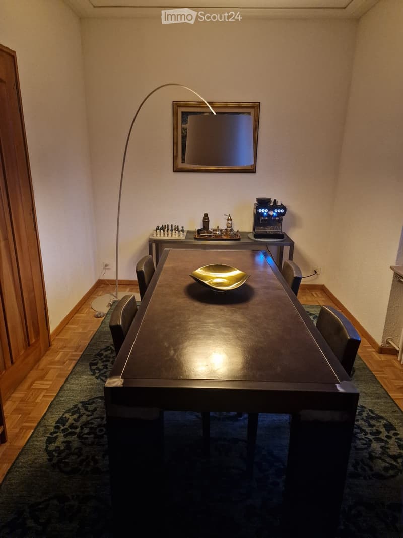 Dining area with a long dark wooden table, four black chairs, a coffee machine, a chessboard, a decorative bowl, a mirror, and a floor lamp.