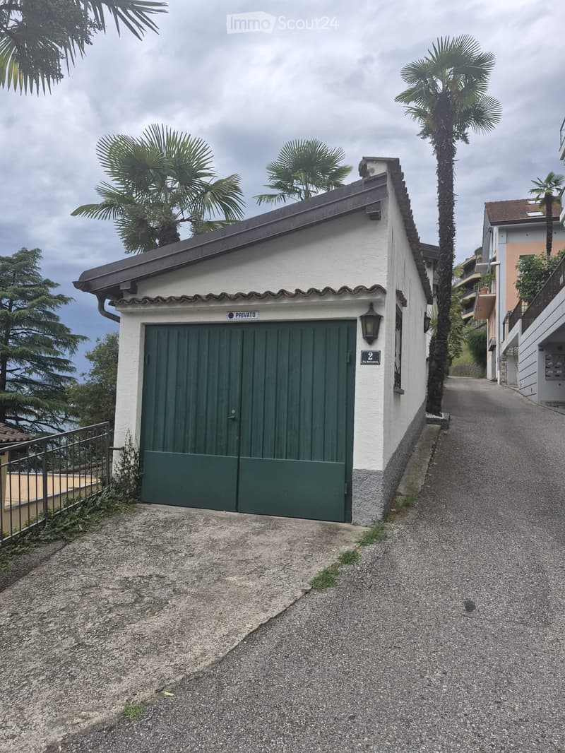 A small, white building with a green garage door, surrounded by palm trees and a gravel driveway.