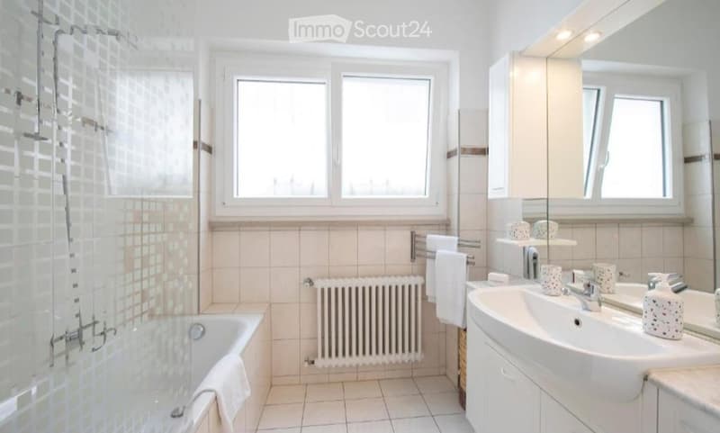Bright bathroom with white tiles, a bathtub, a sink with a mirror, a radiator, and a towel rack. The window offers natural light.