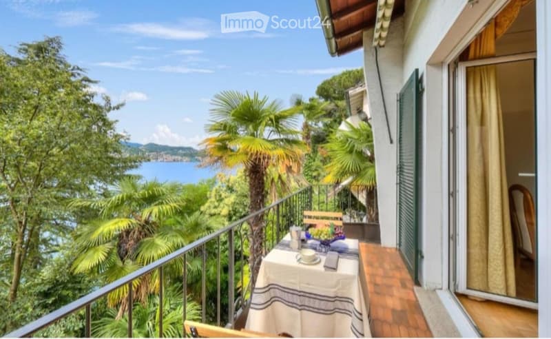 Balcony with a table and chairs, overlooking a scenic lake and lush greenery.