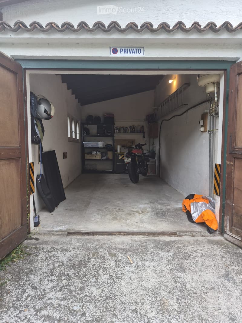 Garage with a motorcycle, helmets, and storage shelves. The garage door is open, revealing a concrete floor and wooden doors on either side.