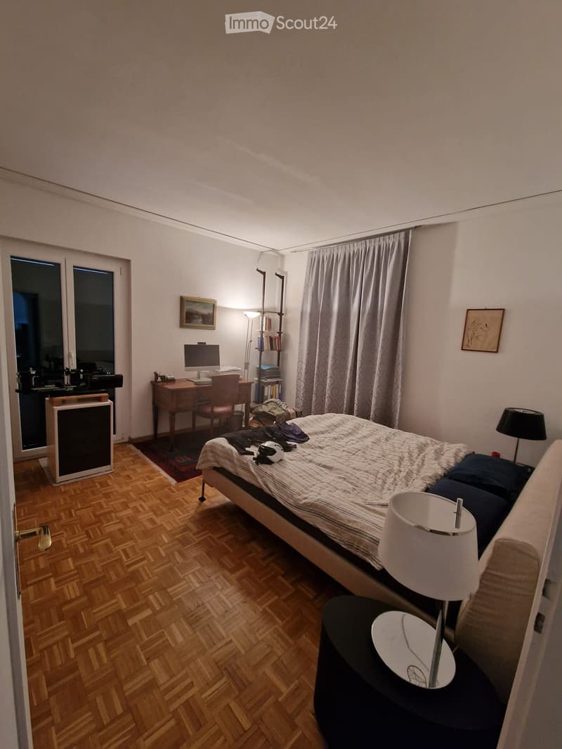 Cozy bedroom with wooden herringbone flooring, a double bed with bedding, a nightstand with a lamp, a desk with a computer monitor, a bookshelf, and a small refrigerator near the window.