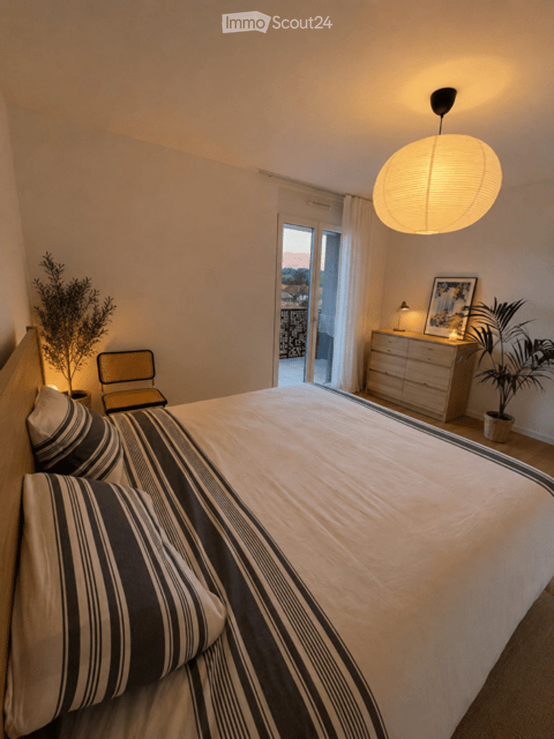 Modern bedroom with a large bed, striped bedding, a nightstand with a lamp, a small chair, and potted plants near the balcony door.
