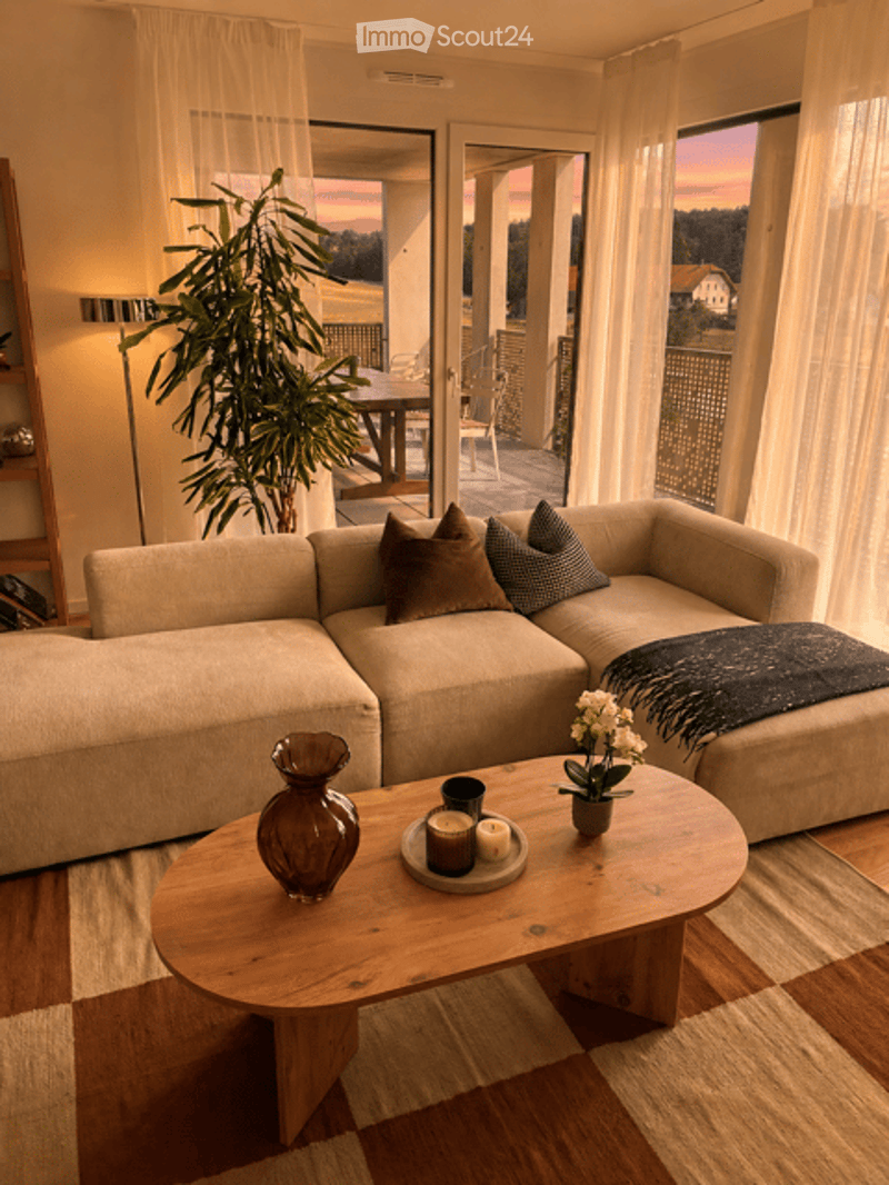 Modern living room with a large sectional sofa, wooden coffee table, decorative vase, candles, and a potted plant. Large windows offer a view and access to a balcony.