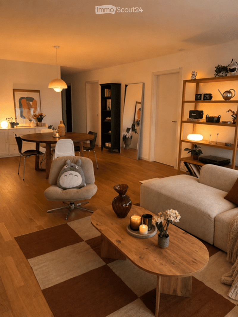 Modern living room with hardwood floors, a grey sofa, a wooden coffee table with candles and a vase, a dining area with a round table and chairs, and a bookshelf with decorative items.