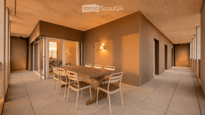 A modern, minimalist dining area with a wooden table and white chairs, surrounded by sleek, contemporary walls and a tiled floor. The space is well-lit with warm wall-mounted lighting, and features large glass doors leading to an adjacent room.