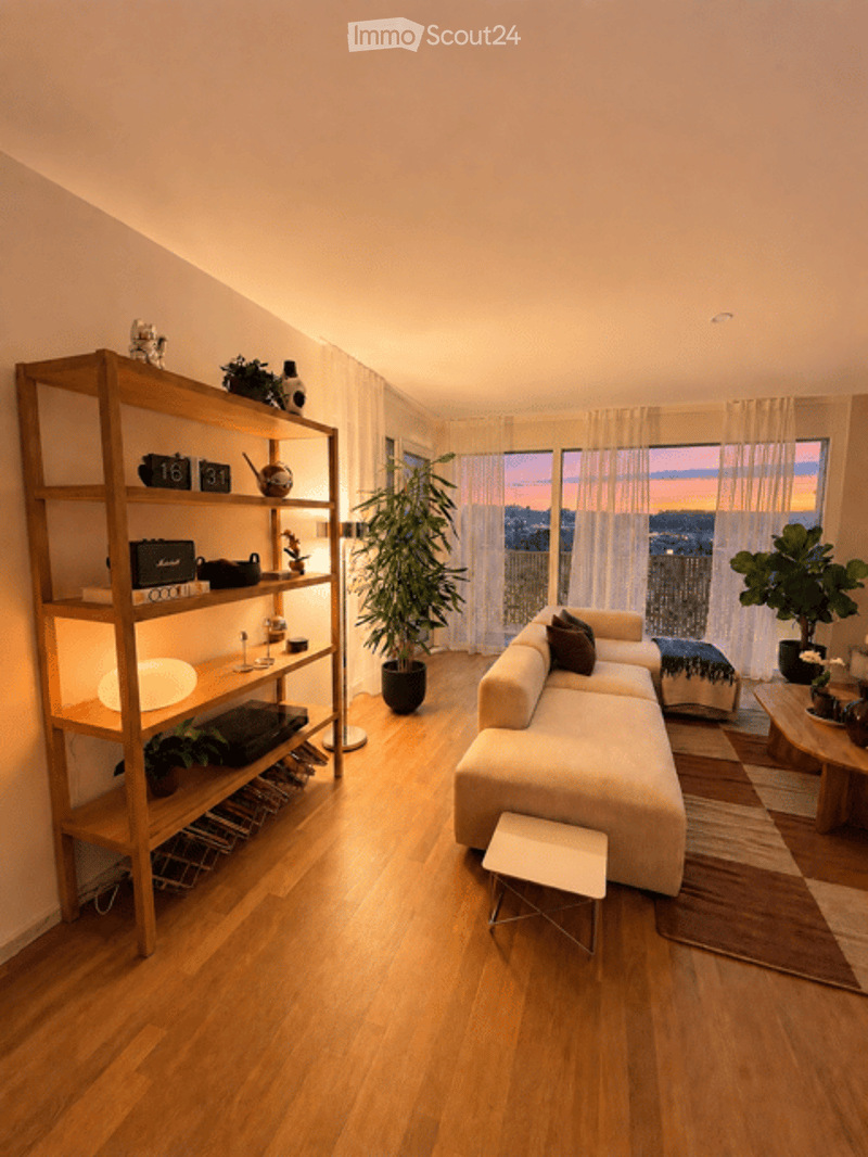 Modern living room with large windows offering a city view, a cream-colored sectional sofa, a wooden shelf with decorative items, a floor lamp, and potted plants.