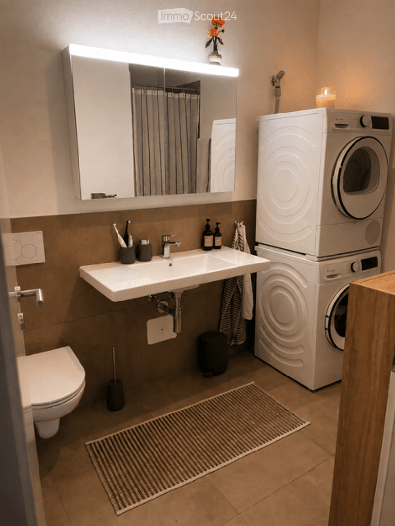 Modern bathroom with a white sink, large mirror, toilet, washing machine, and dryer. Features a towel rack, toothbrush holder, and a small trash bin.