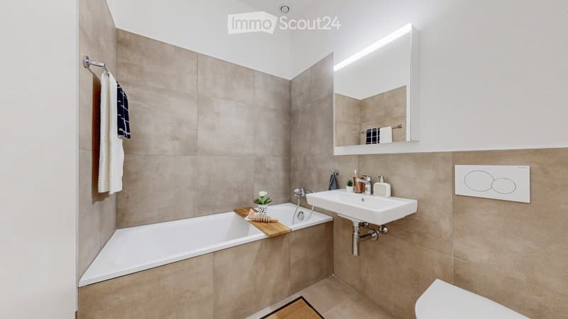 Modern bathroom with beige tiles, a bathtub, a sink, a mirror with built-in lighting, and a toilet. Includes a wooden tray with a small plant and decorative items on the edge of the bathtub.
